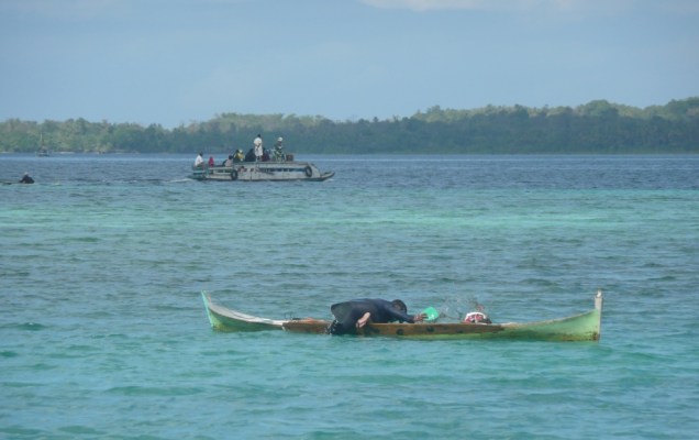 Wanci - Wangi Wangi – Pêcheur. Il est allongé en travers de sa pirogue sans moteur, un pied est chaussé d’une palme pour la propulsion, de la main droite il écope, de la main gauche il tire un filet minuscule et plonge la tête dans l’eau avec un masque pour surveiller le fond.